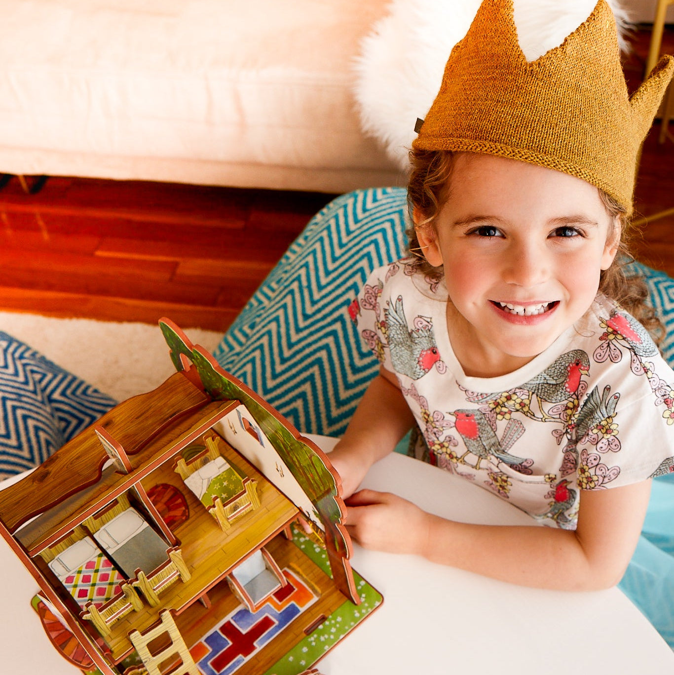 Child wearing a crown holding a toy house, sitting on a bed with a colorful pillow.